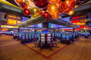 A sprawling casino floor replete with slot machines and other digital games atop a gold and red oriental carpeting and beneath an assortment of red, orange, and gold globular and rectangular lighting fixtures
